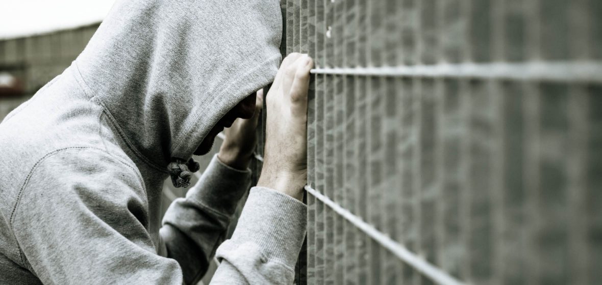 Male wearing hooded sweatshirt leaning against a prison fence