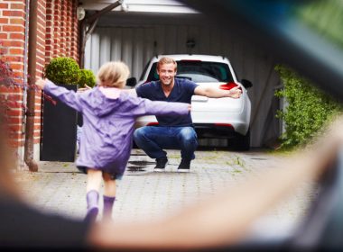 Child runs up a driveway towards a man while a woman sitting in a car watches them.