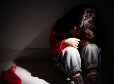 A child buries her head in arms in a dark room.