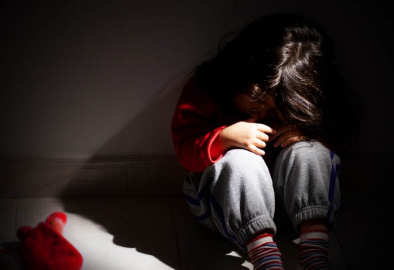 A child buries her head in arms in a dark room.