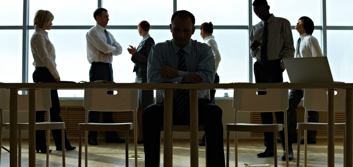 Businessman sits alone at his desk in the shadows while his colleagues stand in the background