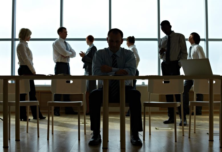 Businessman sits alone at his desk in the shadows while his colleagues stand in the background