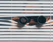 photo of person peering through blinds with binoculars to show surveillance