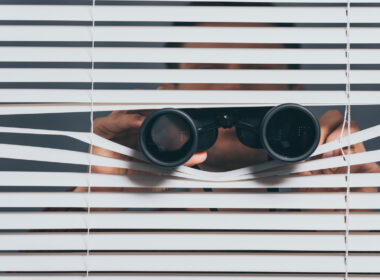 photo of person peering through blinds with binoculars to show surveillance
